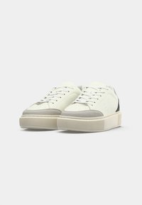 Une paire de baskets basses blanches et grises avec des détails perforés et de grosses semelles beiges sur fond blanc.