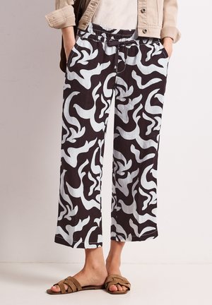 Luźne czarno-białe spodnie z abstrakcyjnym wzorem, z paskiem ściągającym w talii, noszone z jasnobrązowymi sandałami i lekko beżową kurtką.