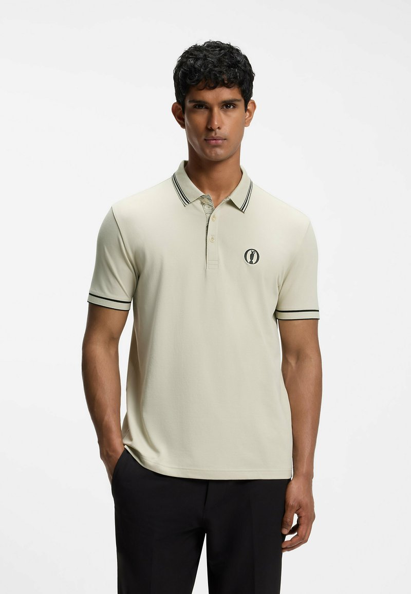 Homme debout, les mains dans les poches, portant un polo beige clair avec des détails noirs et un logo, et un pantalon noir, sur un fond uni.