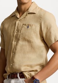 Camisa de lino beige de manga corta con cuello, que presenta un bolsillo en el pecho y un pequeño logo azul. Diseñada con una tela ligera y texturada.