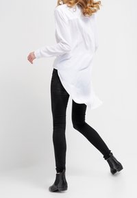 Camisa blanca de manga larga con dobladillo curvado, combinada con jeans ajustados negros y botas de tobillo. Tejido suave, diseño minimalista y silueta ajustada.