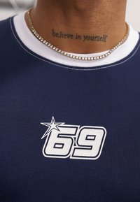 Granatowa bawełniana koszulka z białym okrągłym kołnierzykiem i grafiką "69" z akcentem w postaci gwiazdy. Złoty naszyjnik i widoczny tatuaż na szyi.