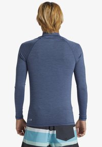 Duge rukava rash guard u tamnoplavoj boji, napravljen od laganog, teksturiranog materijala. Ima raglan rukave i uski kroj, nosi se uz prugaste kratke hlače.