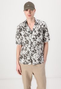 Kratka majica s gumbom s kratkimi rokavi v beli in sivi tie-dye vzorcu; lahkoten material; sproščen kroj z žepom na prsih; kombinirana z bež hlačami.