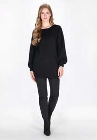 Schwarzer oversized Pullover mit langen, puffigen Ärmeln, rundem Ausschnitt und geripptem Saum, kombiniert mit schmal geschnittenen schwarzen Hosen und hohen Sockenstiefeln.