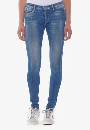 Femme portant un jean bleu slim et des baskets blanches, debout, les bras détendus le long du corps, devant un fond uni.