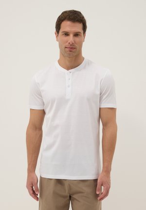 Mann trägt ein schlichtes weißes Henley-Shirt mit kurzen Ärmeln und beige Hose, steht vor einem hellen, neutralen Hintergrund.