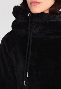 Nainen, joka pukeutunut mustaan karvaiseen huppariin, jossa on kiristysnauhat, näyttää osan kasvoistaan nenästä leuan suuntaan yksiväristä taustaa vasten.