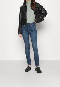 Chaqueta corta de cuero negra, suéter de cuello alto verde claro, jeans ajustados azul oscuro y zapatos sin cordones verde oscuro. Diseño simple, estilo ajustado.