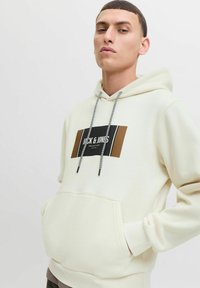 Felpa color crema con tasca a marsupio frontale, patch con logo nero e marrone, e coulisse grigia con un motivo a contrasto.