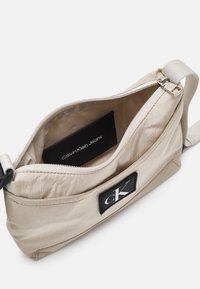 Bolso bandolera de tela beige con cierre de cremallera, que presenta un parche negro con el logo y un bolsillo interior etiquetado como "Calvin Klein Jeans."