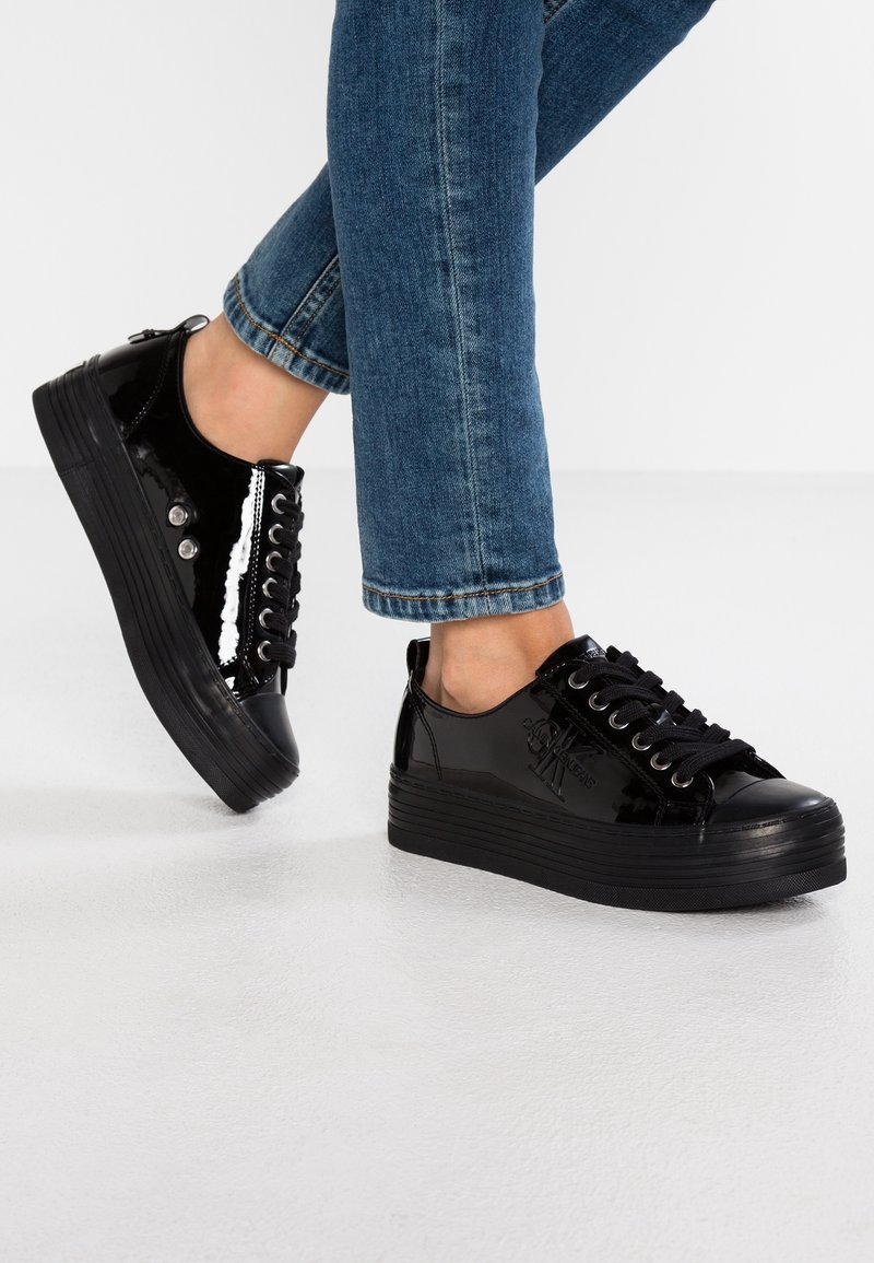 Personne portant des baskets à plateforme noires brillantes avec des lacets noirs et un jean bleu foncé retroussé sur un sol blanc.