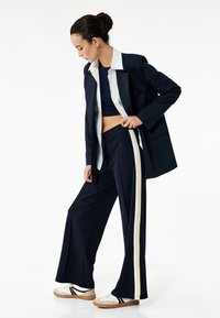 Marineblazer over en marineblå crop top og matchende brede bukser med hvide sidestriber, parret med hvide sneakers og beige detaljer.