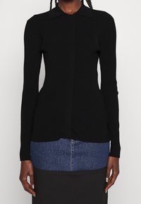 Svart ribbad stickad långärmad topp med krage, knappstängning och en figurnära silhuett. Slät textur med en lätt fall vid fållen.
