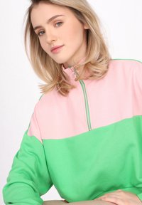 Sudadera con bloque de color que presenta una sección superior en rosa y una sección inferior en verde, con un alto cuello y detalle de cremallera. Tejido suave y texturizado.