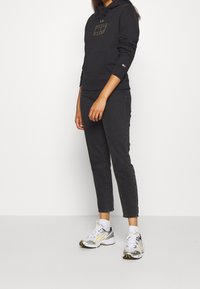 Mujer vestida con sudadera negra, jeans cortos negros, calcetines blancos y zapatillas blancas con detalles en amarillo y negro, de pie contra un fondo claro.