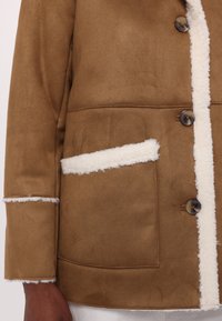 Hnědá bunda s falešným beránkem, s knoflíkovým zapínáním, náprsní kapsou a strukturovaným povrchem.