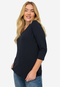 Navyblaues V-Ausschnitt Langarmshirt aus glattem Stoff mit lockerer Passform und abgerundetem Saum. Das Model trägt hellblaue Jeans.