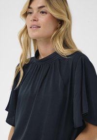 Blouse bleu marine avec des manches courtes et larges. Dotée d'un col haut froncé et d'un tissu doux et lisse avec un léger éclat.