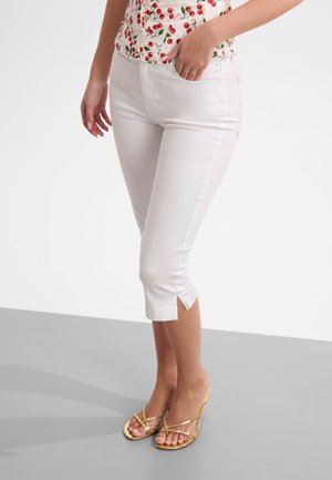 Vrouw draagt witte cropped broek met zijopeningen, een top met kersenprint en gouden sandalen met bandjes, staand op een grijze vloer.