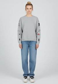 Maglione grigio con design ricamati sulle maniche, abbinato a jeans azzurri chiari e sneakers nere. Design semplice, vestibilità rilassata.