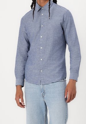 Homme portant une chemise bleu ajustée à manches longues boutonnée, associée à un jean bleu clair, debout devant un fond uni.