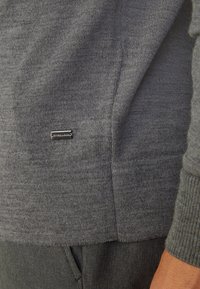 Grauer Strickpullover mit gerippten Bündchen und Saum, ausgestattet mit einem kleinen Metallsignet an der Seite. Weiche Textur, halbschmale Passform.