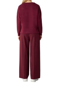 Vrouw in een losvallende wijnrode sweatshirt en wijde broek, staand met de rug naar de kijker, witte sneakers zichtbaar.