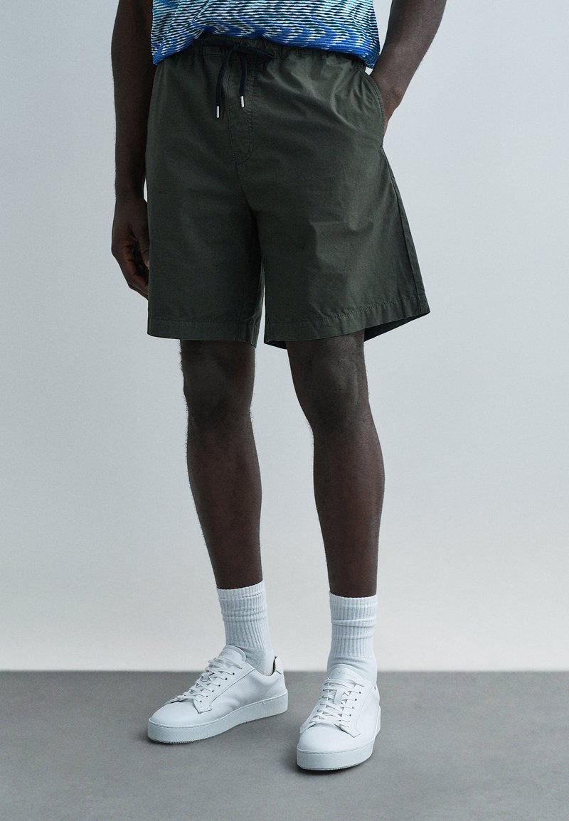 Olivově zelené šortky z lehké látky s elastickým pásem. Kombinované s bílými teniskami a bílými ponožkami po kotníky.