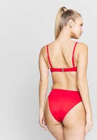 Piros bikini szett állítható pántokkal, aranyszínű csattal a hátsó részen, magasan szabott alsó résszel és sima textúrájú anyaggal.