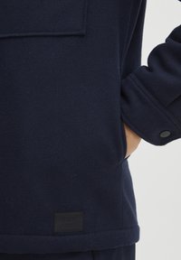 Jachetă din lână navy, cu un buzunar pe piept, manșete cu nasturi și o etichetă de stofă rectangulară la tiv. Textură moale și design adaptat.