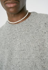 Grauer Strickpullover mit einer strukturierten Oberfläche, die kleine Punkte aufweist. Akzentuiert mit einer einfachen Perlenkette um den Hals.