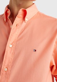 Nahaufnahme einer Person, die ein pfirsichfarbenes Hemd mit Knöpfen und einem kleinen rechteckigen Logo auf der Brust trägt.