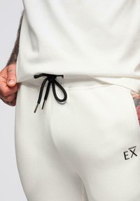 Białe spodnie dresowe z czarnym sznurkiem, metalowymi końcówkami i haftowanym logo "EX" na lewym biodrze. Miękka tkanina i dopasowany krój.