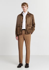 Giacca in suede marrone con bottoni e due tasche frontali, abbinata a pantaloni beige e scarpe scure. Camicia bianca indossata sotto.