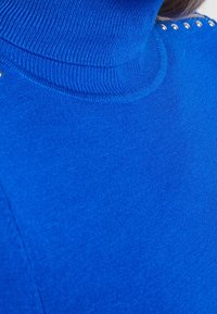 Blauer Rollkragenpullover aus Strickmaterial, mit glatter Textur und metallischen Akzentnieten entlang der Schulternaht.