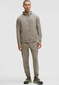 Lichte tan kleurige jas met een capuchon, rits aan de voorkant en twee zijzakken, gecombineerd met bijpassende broek en bruin-groene sneakers. Geweven stof.