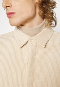Chemise en velours côtelé beige avec une texture ribbed, dotée d'un col classique et d'une fermeture à un bouton. Un col roulé crème est visible en dessous.