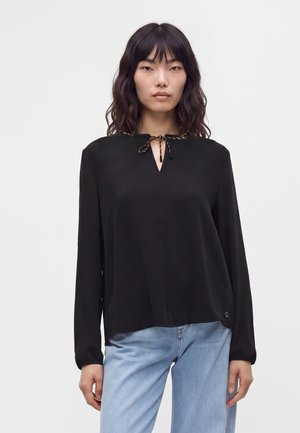 Femme aux cheveux ondulés mi-longs porte un chemisier noir ample à manches longues avec un col à nouer et un jean bleu clair sur un fond uni.