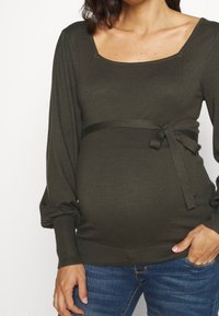 Maglione premaman verde oliva con scollatura quadrata, maniche a sbuffo e un nastrino di tessuto in vita. Realizzato in un morbido materiale a costine.