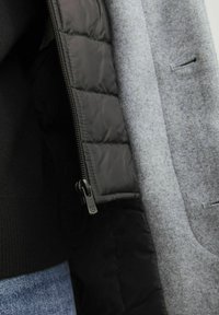 Chaqueta de mezcla de lana gris con un forro interior acolchado negro, que presenta una cremallera y sutiles detalles de costura. Primer plano de la tela en capas.