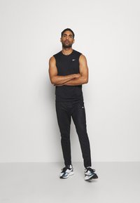 Camiseta preta sem mangas, calças de desporto pretas e ténis brancos com detalhes em teal. Modelo está de braços cruzados contra um fundo branco.
