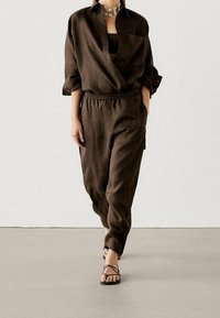 Donkerbruine linnen shirt met opgerolde mouwen, in combinatie met bijpassende losse broeken met een elastische tailleband, en zwarte sandaaltjes met bandjes.