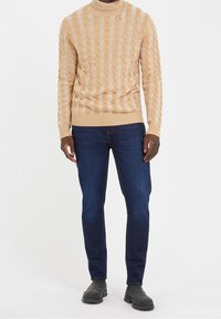 Béžový svetr s kabelovým vzorem a vysokým límcem, v kombinaci s tmavě modrými úzkými džínami a šedými kotníkovými botami. Jsou viditelné texturované vzory tkaniny.