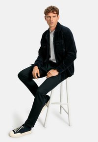 Giacca in velluto a coste nera con due tasche sul petto, indossata sopra una polo bianca. Pantaloni neri slim-fit e sneakers blu navy con suola cremoso.