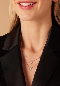 Zilverkleurige gelaagde kettingen met een maan- en sterpendant, gedragen over een zwart blouse met kraag, wat een strakke uitstraling benadrukt.