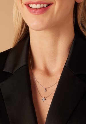 Zilverkleurige gelaagde kettingen met een maan- en sterpendant, gedragen over een zwart blouse met kraag, wat een strakke uitstraling benadrukt.
