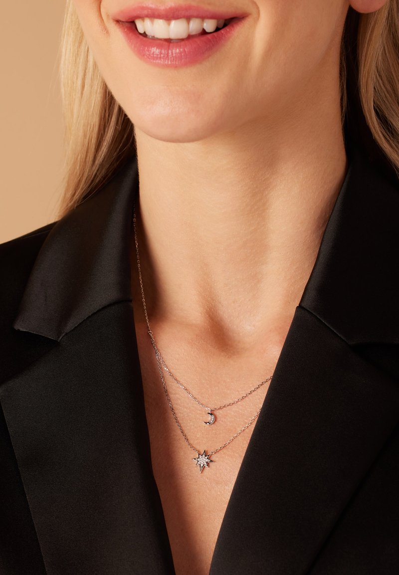 Zilverkleurige gelaagde kettingen met een maan- en sterpendant, gedragen over een zwart blouse met kraag, wat een strakke uitstraling benadrukt.