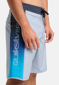 Svetlo šedé šortky na plávanie s modrým gradientovým vzorom a čiernymi akcentmi. Majú pás s šnúrkou a logo na boku.