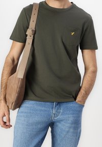 Homme portant un t-shirt vert olive avec un petit logo d’oiseau jaune sur la poche, un jean bleu clair et portant un sac à bandoulière beige.